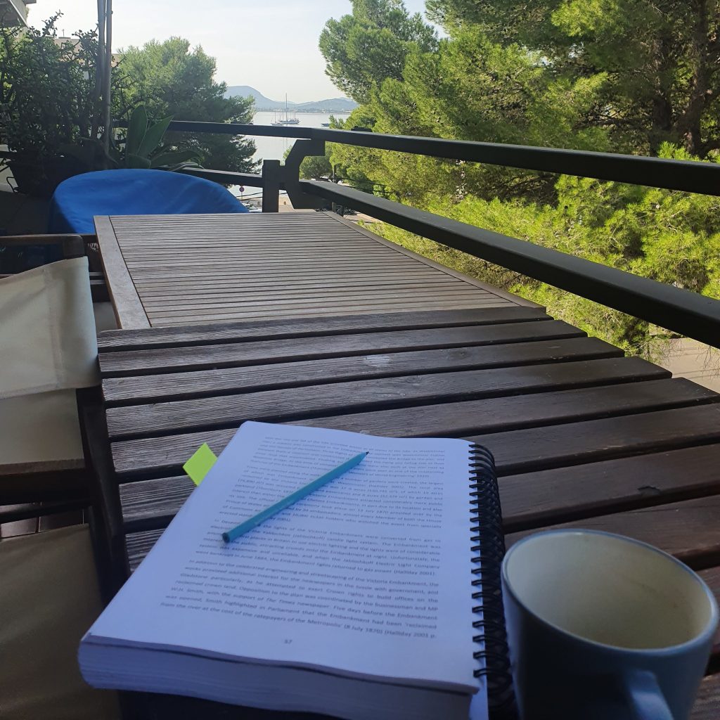 My thesis sits open at page 57 on a balcony table, with a pencil on top and a coffee cup beside. The view from the balcony between the trees is of the sea at Port de Pollenca, Mallorca where a couple of sailing yachts are moored up, and the hills in the distance. 
